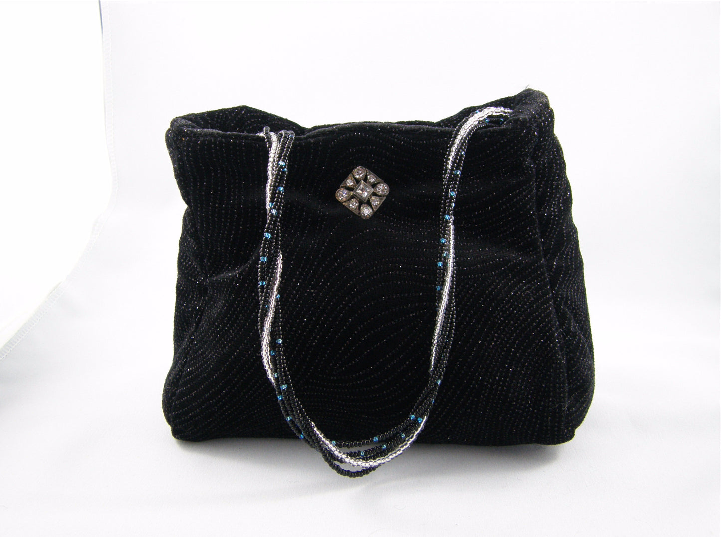 Black and Silver Evening Bag from Jenny Jag-Wear Design. The photo shows this compact clutch in black holiday fabric with silver glitter. The beaded handle is hand crafted from silver, black and turquoise seed beads, and embellished with a small crystal brooch. The Merle Collection from Jenny Jag-Wear Design